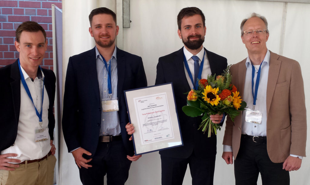 Vier teils bärtige Herren, von denen einer eine Urkunde und einen Blumenstrauss trägt; er ist Preisträger des Young Professionals‘ Award Logistics.