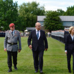 Ein Soldat in Uniform, ein Mann in einem dunklen anzug und eine Frau in einem blauen Kostüm gehen vor einer Reihe angetretener Soldaten längs.