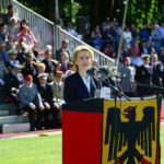 Eine blonde Dame im blauen Kostüm steht hinter einem Rednerpult, das in eine Bundesdienstflagge eingehült ist, dahinter sitzen Männer in bunten Uniformen auf einer Tribüne.