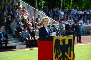 Eine blonde Dame im blauen Kostüm steht hinter einem Rednerpult, das in eine Bundesdienstflagge eingehült ist, dahinter sitzen Männer in bunten Uniformen auf einer Tribüne.
