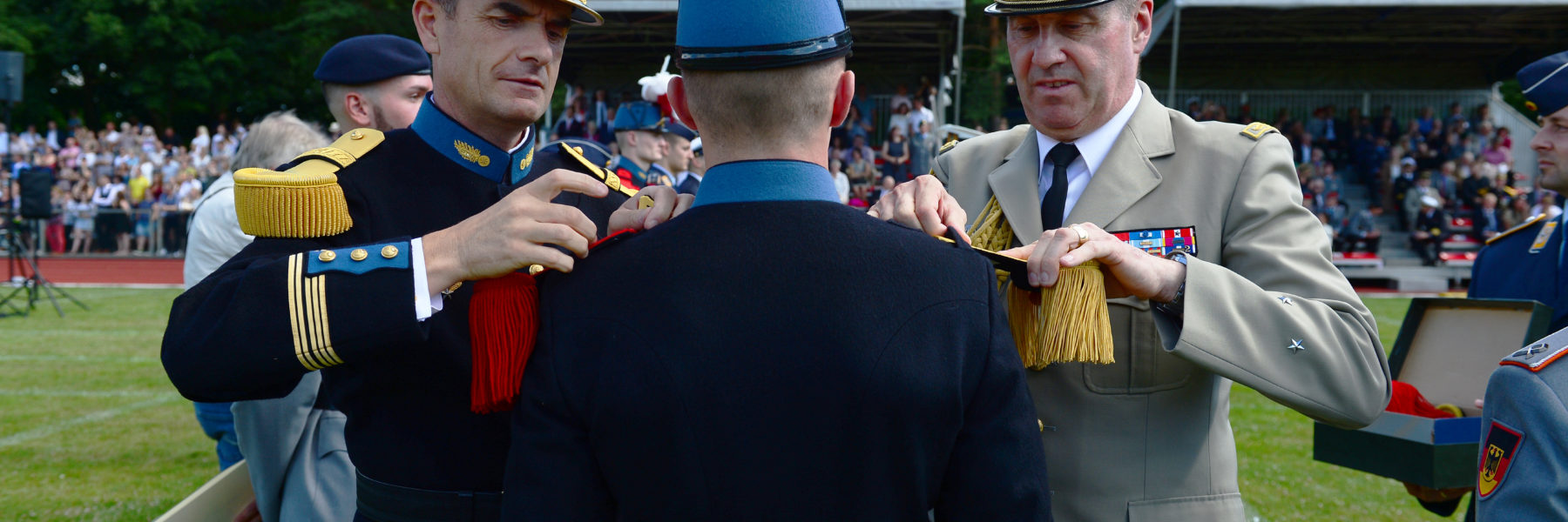 Zwei Soldaten in bunten Uniformen stecken einem dritten Soldaten mit Federbusch am Hut neue Epauletten auf die Schulter.