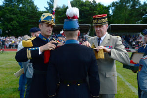 Zwei Soldaten in bunten Uniformen stecken einem dritten Soldaten mit Federbusch am Hut neue Epauletten auf die Schulter.