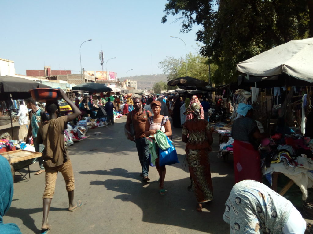 Dunkelhäutige Menschen tragen Dinge vor einer fremd anmutenden Marktplatzkulisse hin und her.