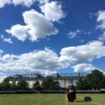 Ein Soldat in Uniform am Rednerpult vor einer in enorm langer Reihe angetretenen Formation von Soldaten auf einem grünen Rasen, dahinter blauer Himmel mit Schäfchenwolken.