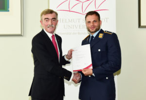 Ein älterer Mann in einem dunklen Anzug überreicht einem jüngeren Mann in Luftwaffenuniform eine Urkunde.