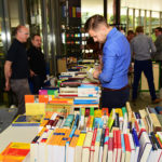 Ein junger Mann im blauen Hemd schaut auf einen großen Bücherstapel.