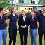 Eine blonde Dame im blauen Kostüm zwischen vier jungen leuten in blauen Polohemden mit der Aufschrift Bundeswehr, dahinter zwei Pferde.