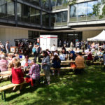 Viele Menschen sitzen im Schatten eines Baumes an Bierzeltgarnituren.