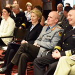 Frauen in Kostümen und Männer in Uniformen sitzen in einem Saal voller Menschen.