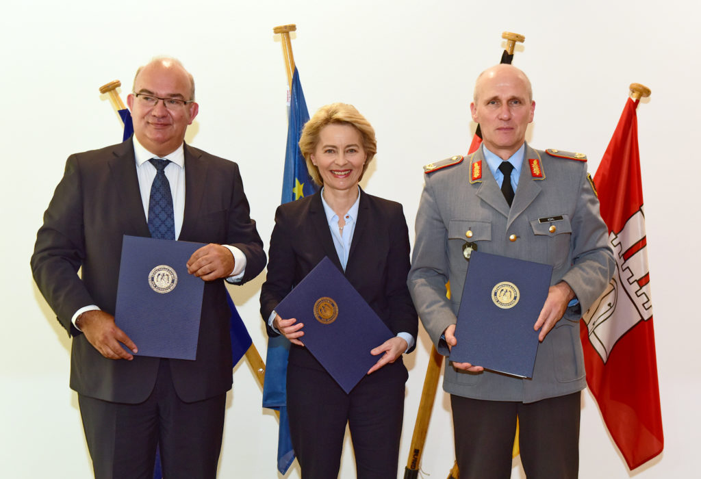 Ein Mann in einem Dunklen Anzug, eine Frau im dunklen Kostüm und ein Soldat in Generalsuniform halten Blaue Dokumentenmappen mit einem goldfarbenen Prägewappen.