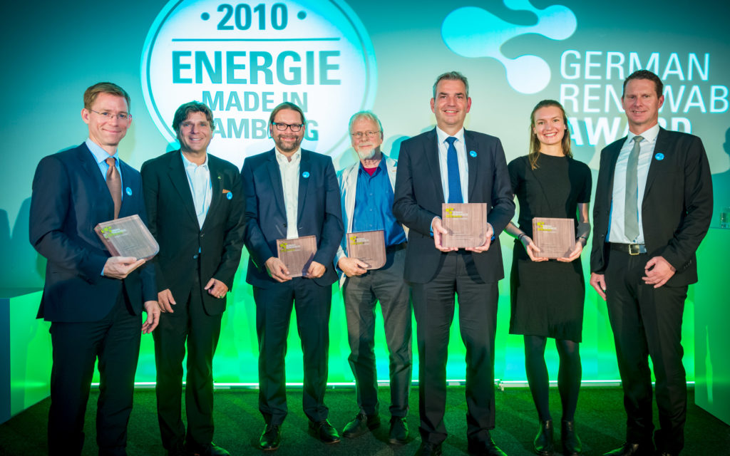 7 Menschen stehen auf einer Bühne, darunter 6 Männer und nur eine Frau. Die Frau ist die Hauptperson. Sie hält eine Tafel, auf der German Renewables Award 2018 steht.