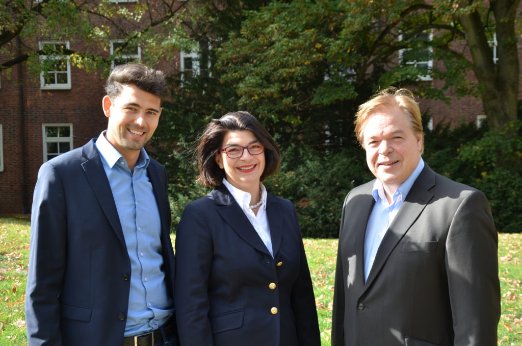 Ein jüngerer Mann in einem blauen Sakko, eine Frau in einem blauen Kostüm und ein etwas älterer Mann in grauem Sakko blinzeln in die Sonne.