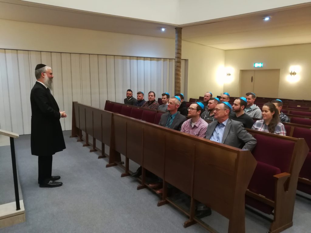 Ein Rabbiner spricht zu einer Gruppe von Menschen, von denen die männlichen alle eine blaue Kipa tragen