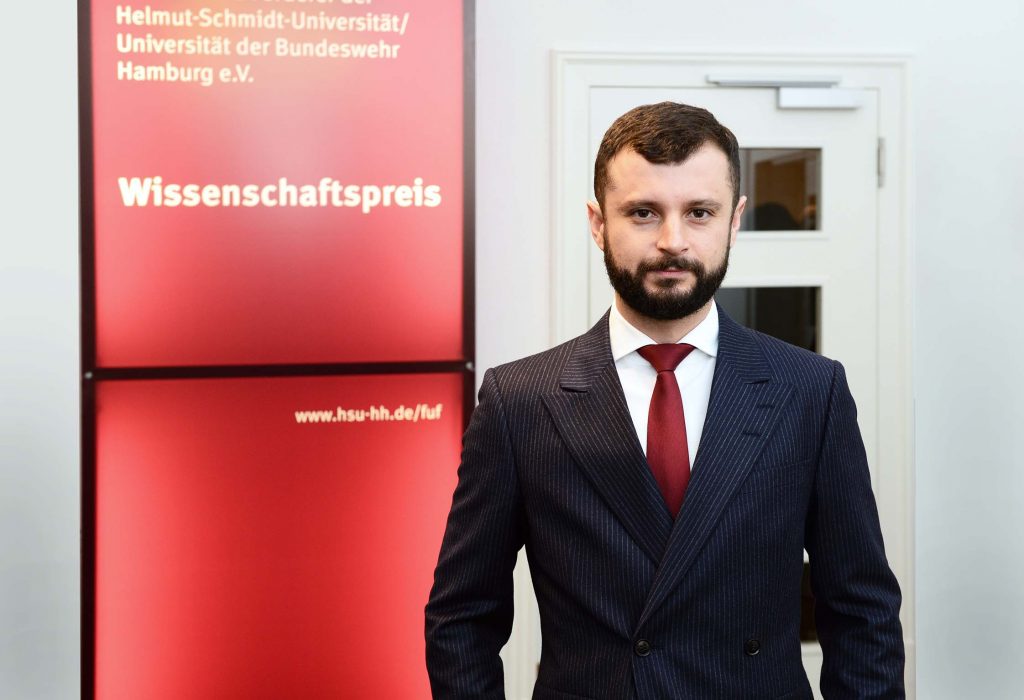 Ein bärtiger junger Mann im blauen Zweireiher mit Nadelstreifen vor einer Tafel mit der Aufschrift Wissenschaftspreis