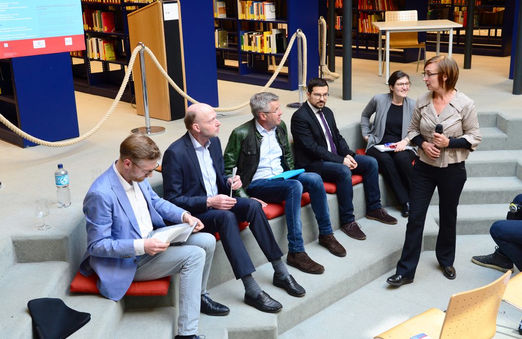 Eine Reihe von Menschen sitzen nebeneinander in einer Bibliothek, eine Frau mit einem Mikrofon steht davor.