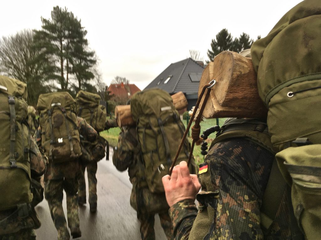 Soldaten im Feldanzug mit Rucksäcken, einige tragen zusätzlich Meterhölzer im Nacken