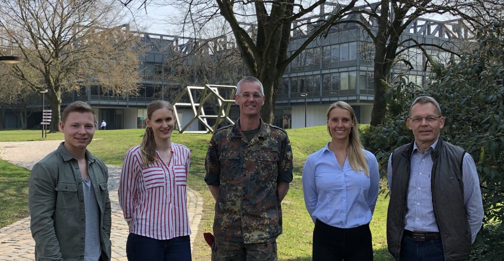 Fünf Menschen, davon zwei Frauen und drei Männer, einer davon im Kampfanzug der Bundeswehr, stehen nebeneinander und haben die Hänbde hinterm Rücken verschränkt.