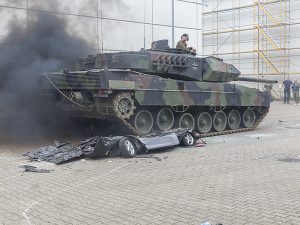 Ein Panzer hat ein Auto überfahren. Das Auto ist platt, der Panzer qualmt