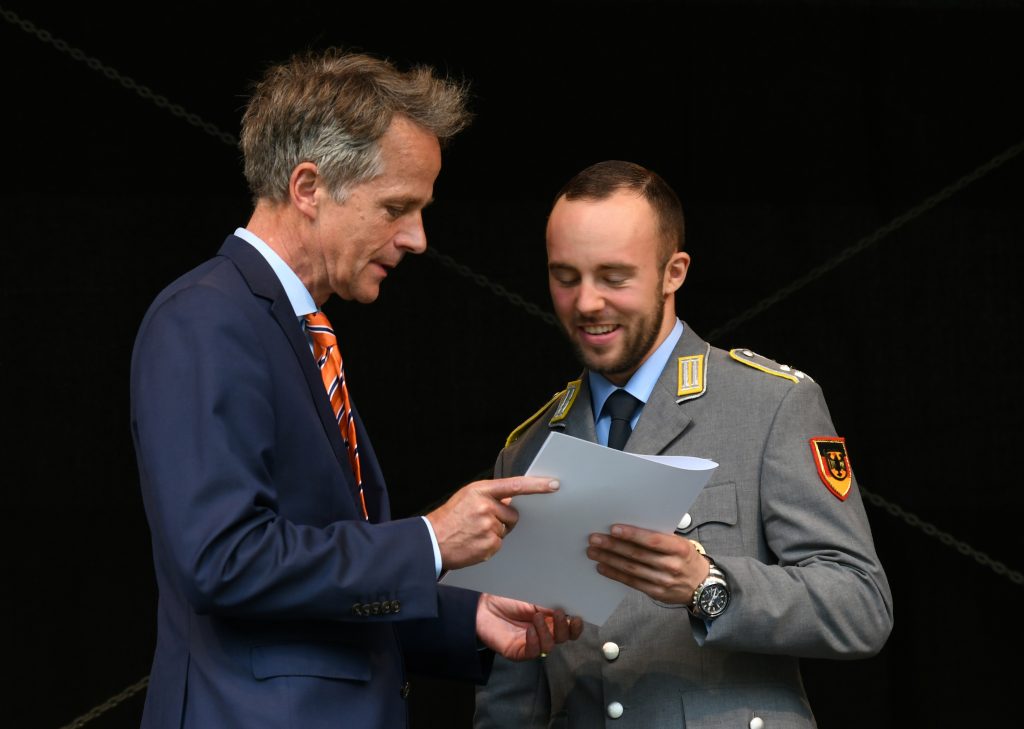 Ein Mann im blauen Anzug überreicht einem Soldaten in Heeresuniform eine Urkunde