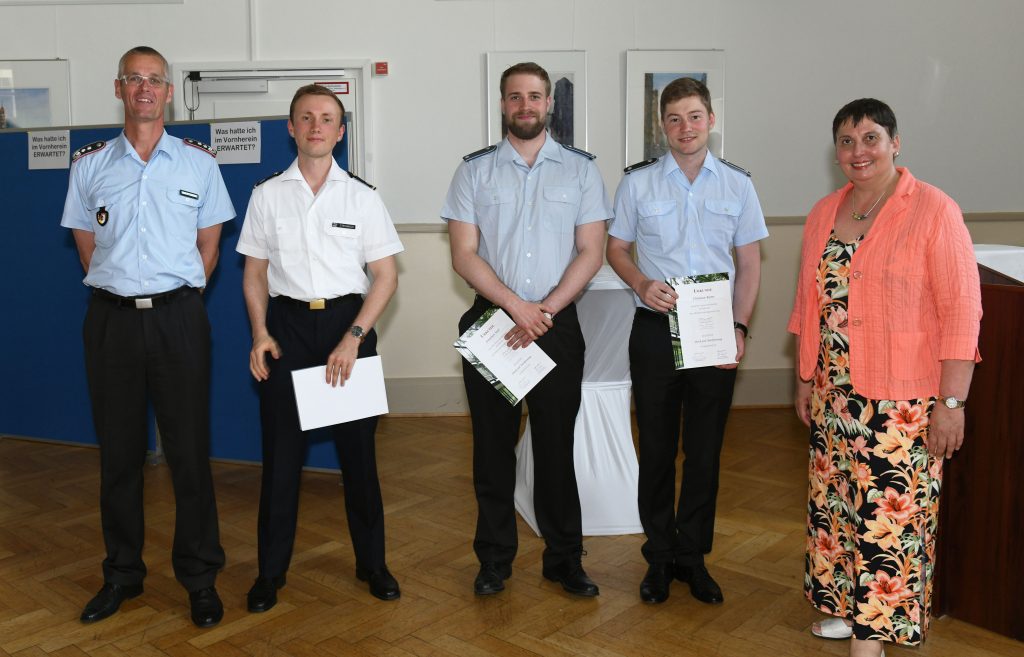 Einige Soldaten in Uniform halten Urkunden in den Händen, daneben steht eine Frau in einem bunten Kleid