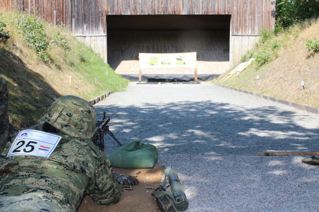 Ein Soldat liegt auf dem Boden und schießt mit einem Gewehr auf eine Zeilscheibe, dabei trägt er einen Helm. Auf dem Rücken hat er ein Schild mit einer Wettkampfnummer