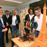 Fünf Männer und eine Frau stehen um einen 3D-Drucker. Im Vordergrund ist ein bereits gedruckter Eiffelturm aus orangenem Kunststoff zu sehen.