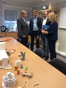 Drei Herren in dunklen Anzügen erklären einer blonden Frau ein Objekt, das von einem 3D-Drucker gedruckt wurde.