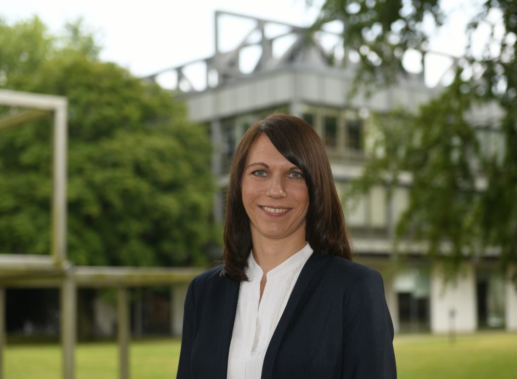 Eine Frau mit offenem Haar steht zwischen zwei Bücherregalen. sie trägt eine weiße Bluse und ein dunkles Jackett vor dem Universitätsgebäude. Sie lächelt.