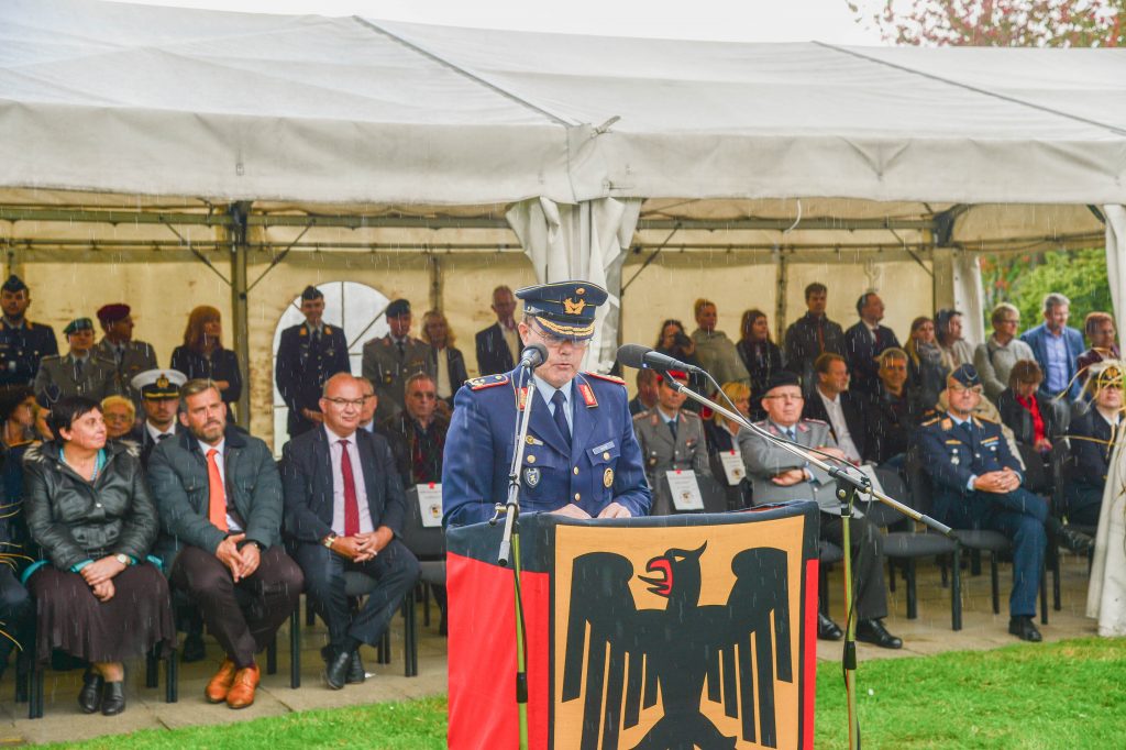 Ein Soldat steht an einem Rednerpult, im Hintergrund ist ein Zelt zu sehen, in dem viele Leute sitzen