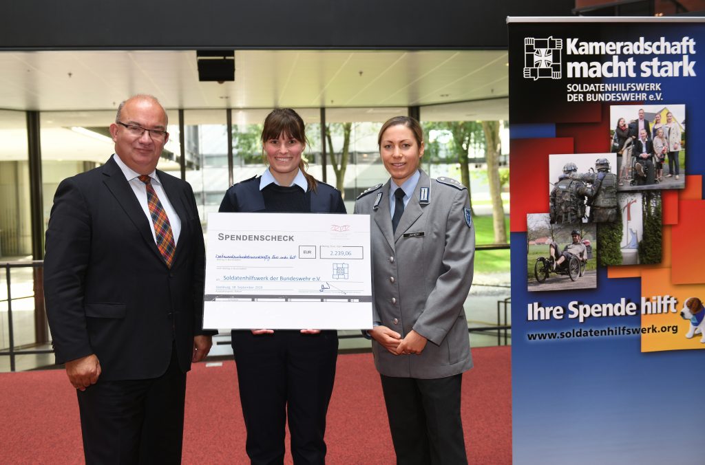 Ein Mann in einem dunklen Anzug mit karierter Krawatte und zwei Soldatinnen in Uniform, von denen eine einen übergroßen Barscheck über 2.239,06 Euro hält.