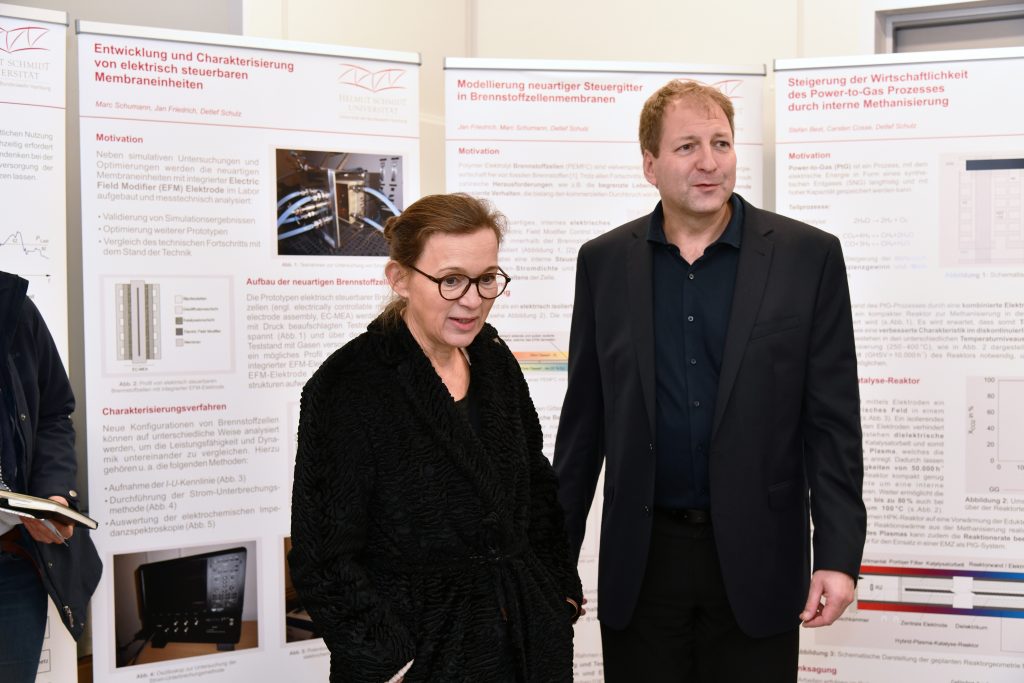 Eine Dame in einem schwarzen Mantel und ein Mann im dunklen Anzug vor einer Reihe von Postern