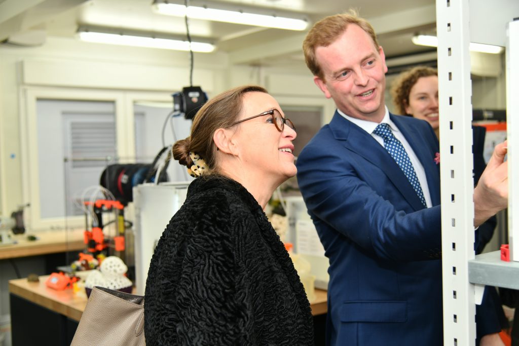 Eine Dame in einem schwarzen Mantel lässt sich von einem jungen Mann im blauen Sakko etwas erklären.