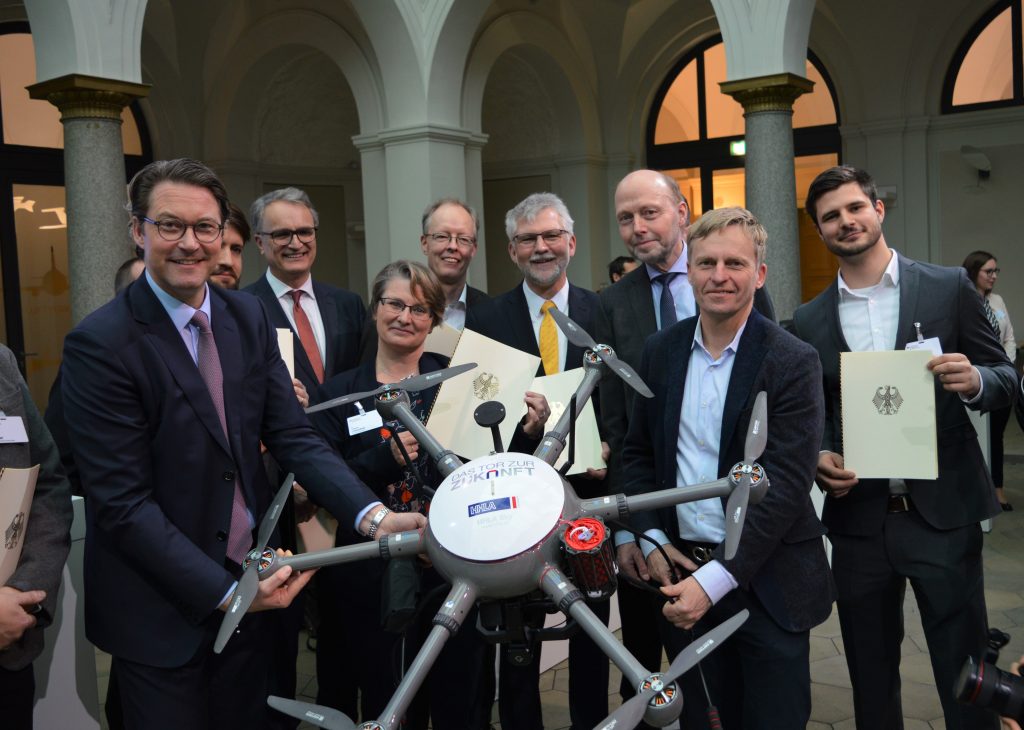 Ach Männer und eine Frau, zwei von Ihnen halten eine Drohne, die restlichen halten Urkunden mit dem Bundesadler