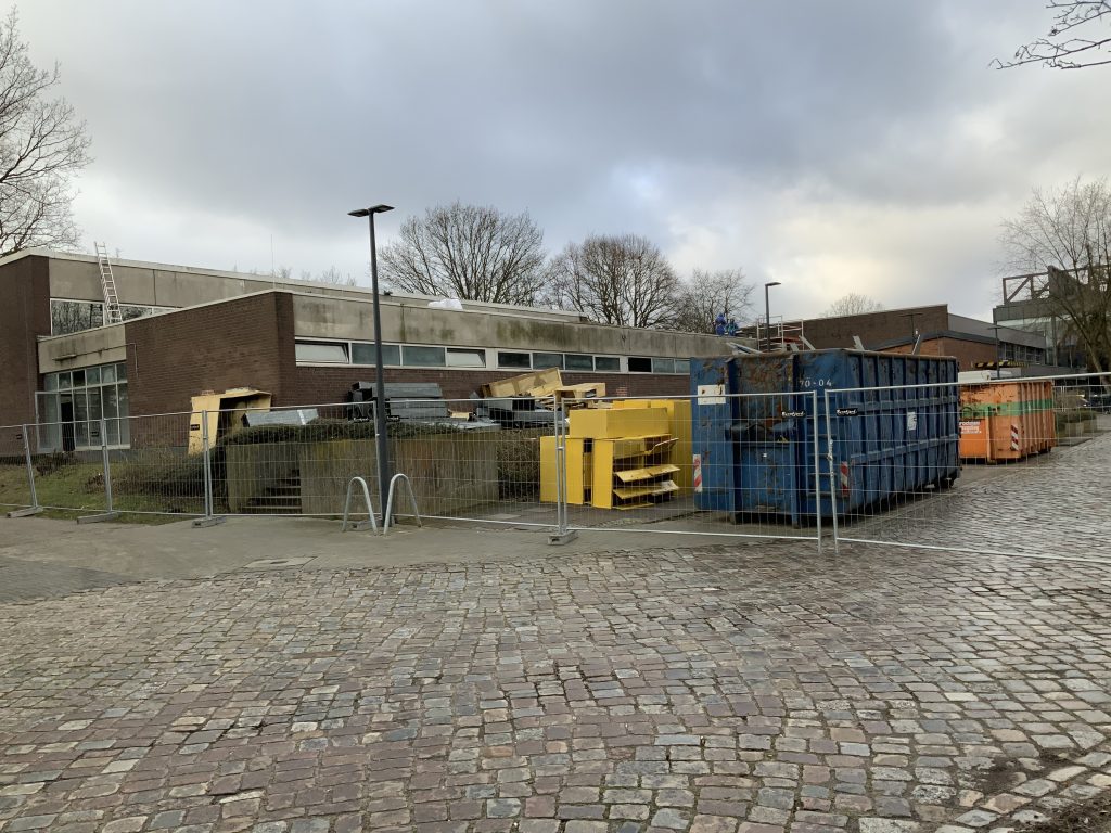 Container und Schrott stehen hinter einem bauzaun und vor einem Gebäude