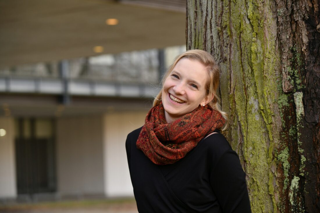 Eine blonde junge Frau, die an einem Baum lehnt. Im Hintergrund das Universitätsgebäude.