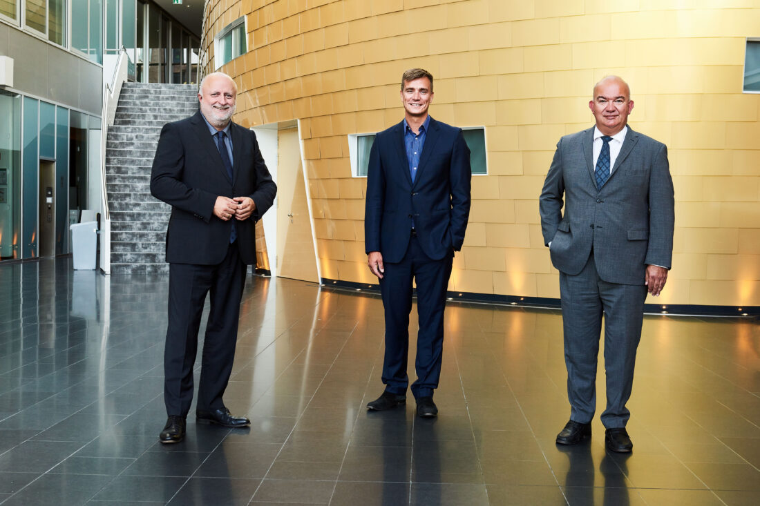 Drei Herren in Anzügen stehen vor einer futuristisch anmutenden Wand