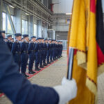 Soldaten in Luftwaffenuniform, davor eine Faust mit weißem Handsschuh, die die Bundesdienstflagge hält.