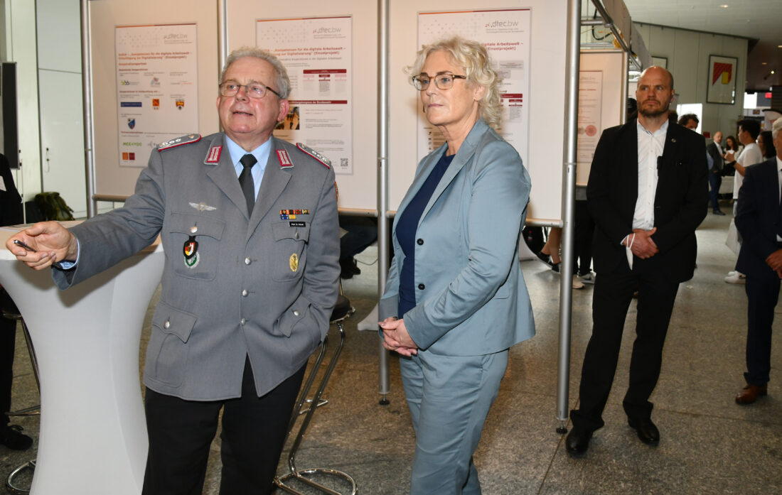 Ein Offizier in Heeresuniform und eine Frau in einem taubenblauen Kostüm stehen vor einer Posterwand