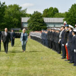 Drei Männer, zwei davon in Uniform, und eine blonde Frau in einem taubenblauen Kostüm schreiten die front einer angetretenen Formation von Soldaten ab.