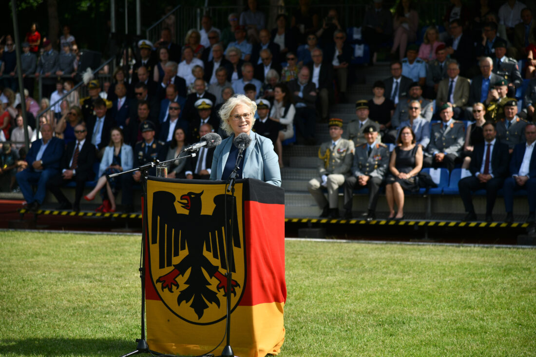 Eine blonde Frau in einem taubenblauen Kostüm steht hinter einem Rednerpult, das in eine Bundedienstflagge eingepackt ist.