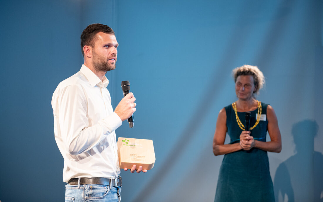 Ein Mann im weißen Hemd mit Mikrofon in der einen und einem Holzobjekt in der anderen Hand