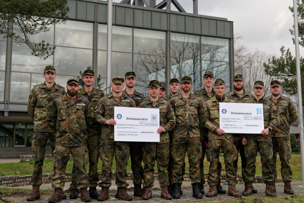 Studierende in Bundeswehr-Tarnuniformen präsentieren zwei Spendenschecks aus den Erlösen des Benfiz-Weihnachtsmarkts 2023 an der Helmut-Schmidt-Universität/Universität der Bundeswehr Hamburg.