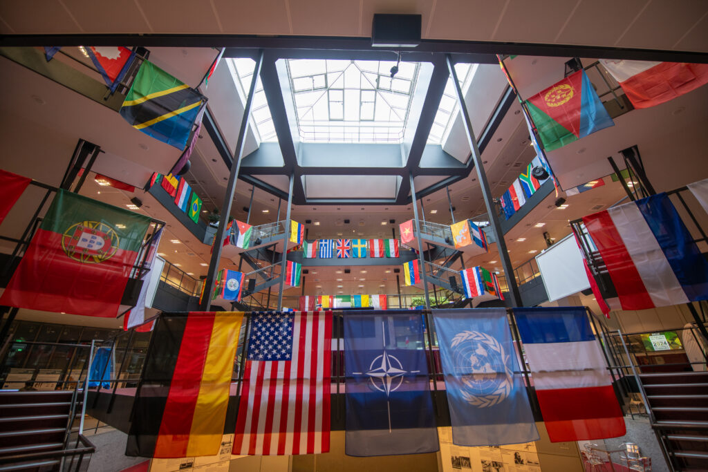Innenansicht eines mehrstöckigen Gebäudes mit offener Architektur, indem zahlreiche internationale Flaggen auf mehreren Ebenen hängen. Darunter die Flagge Deutschlands, der USA, der NATO und den Vereinten Nationen.