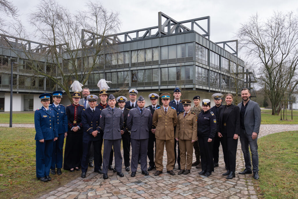 Gruppe internationaler Militärangehöriger in unterschiedlichen Uniformen steht auf einem Campusgelände vor dem Universitätsgebäude der Helmut-Schmidt-Universität/Universität der Bundeswehr.