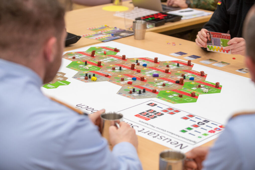 Teilnehmende führen ein Wargaming‑Szenario an einem Tisch mit Spielplan, Spielfiguren und Karten durch.