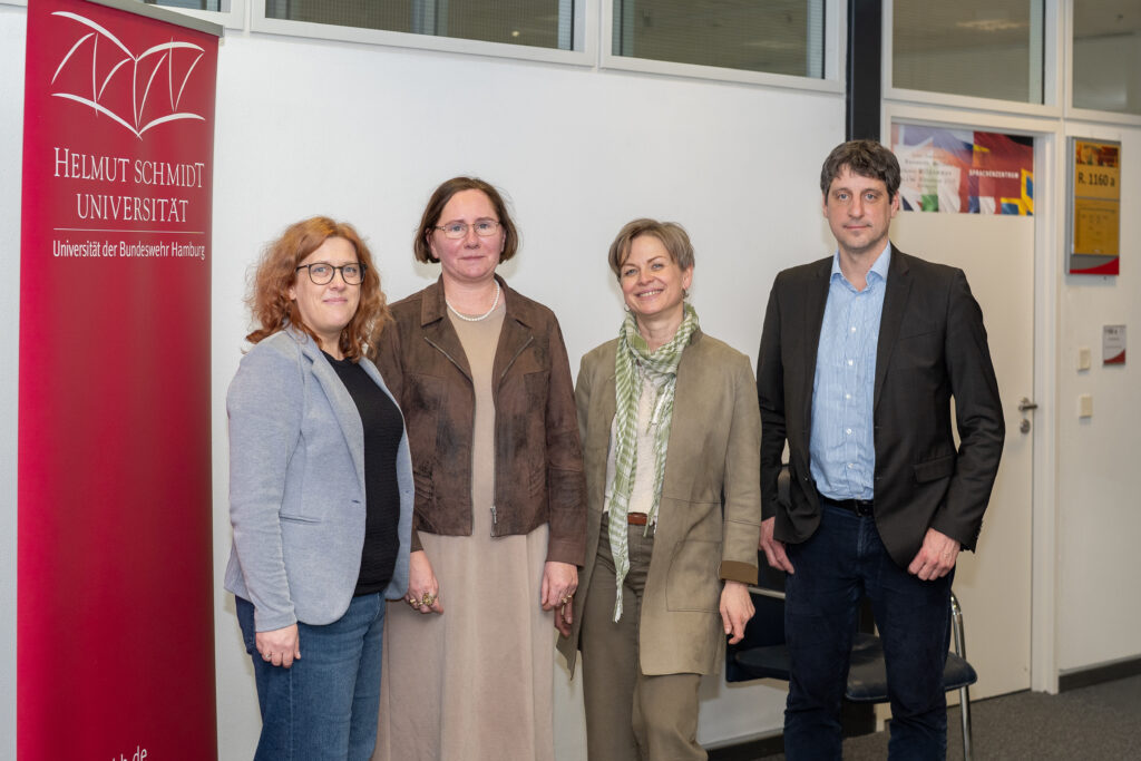 Vier Personen stehen in einem Universitätsgebäude neben einem Roll‑up der Helmut‑Schmidt‑Universität/Universität der Bundeswehr Hamburg.