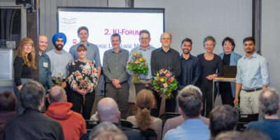 Gruppenfoto von Teilnehmenden des 2. KI‑Forums vor einer Präsentationsfolie in einem Veranstaltungsraum.
