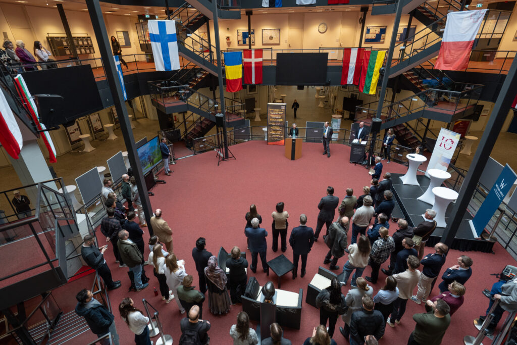 Publikum steht im Rund um eine zentrale Veranstaltungsfläche in einem großen, offenen Lichthof.