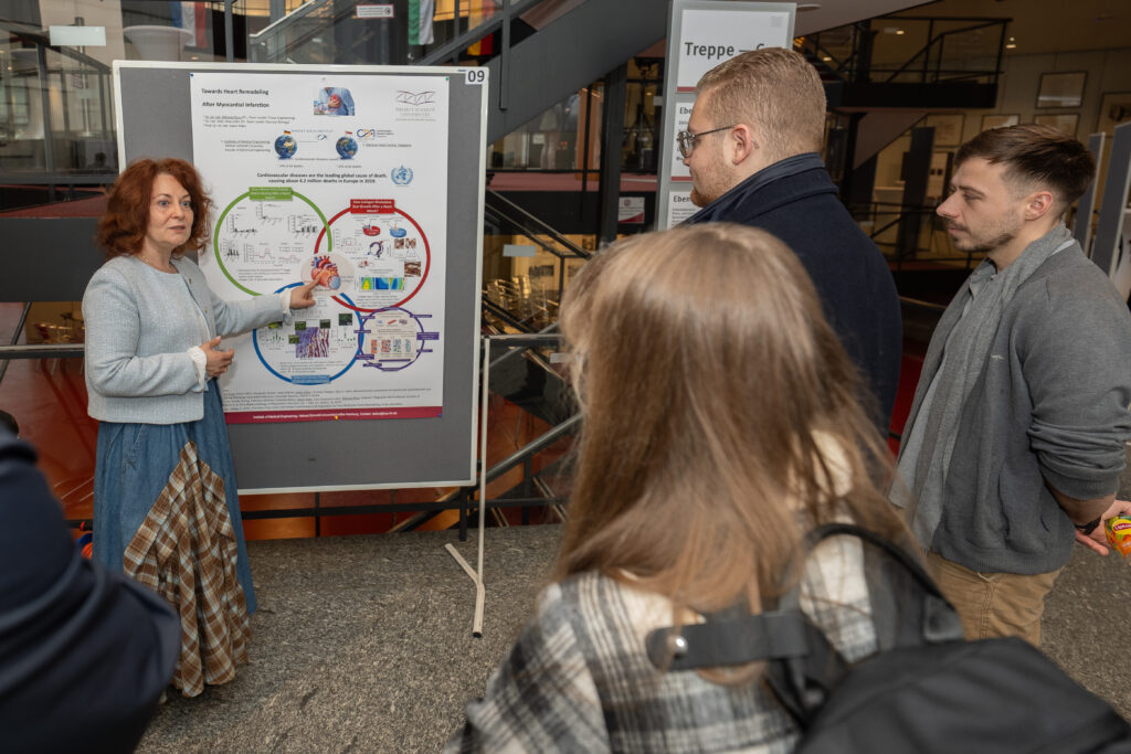 Eine Frau erklärt ein wissenschaftliches Poster vor einer kleinen Gruppe in einem offenen Foyer.