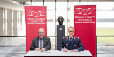 Zwei Männer sitzen an einem Tisch vor einer Helmut-Schmidt-Büste und unterzeichnen einen Vertrag.